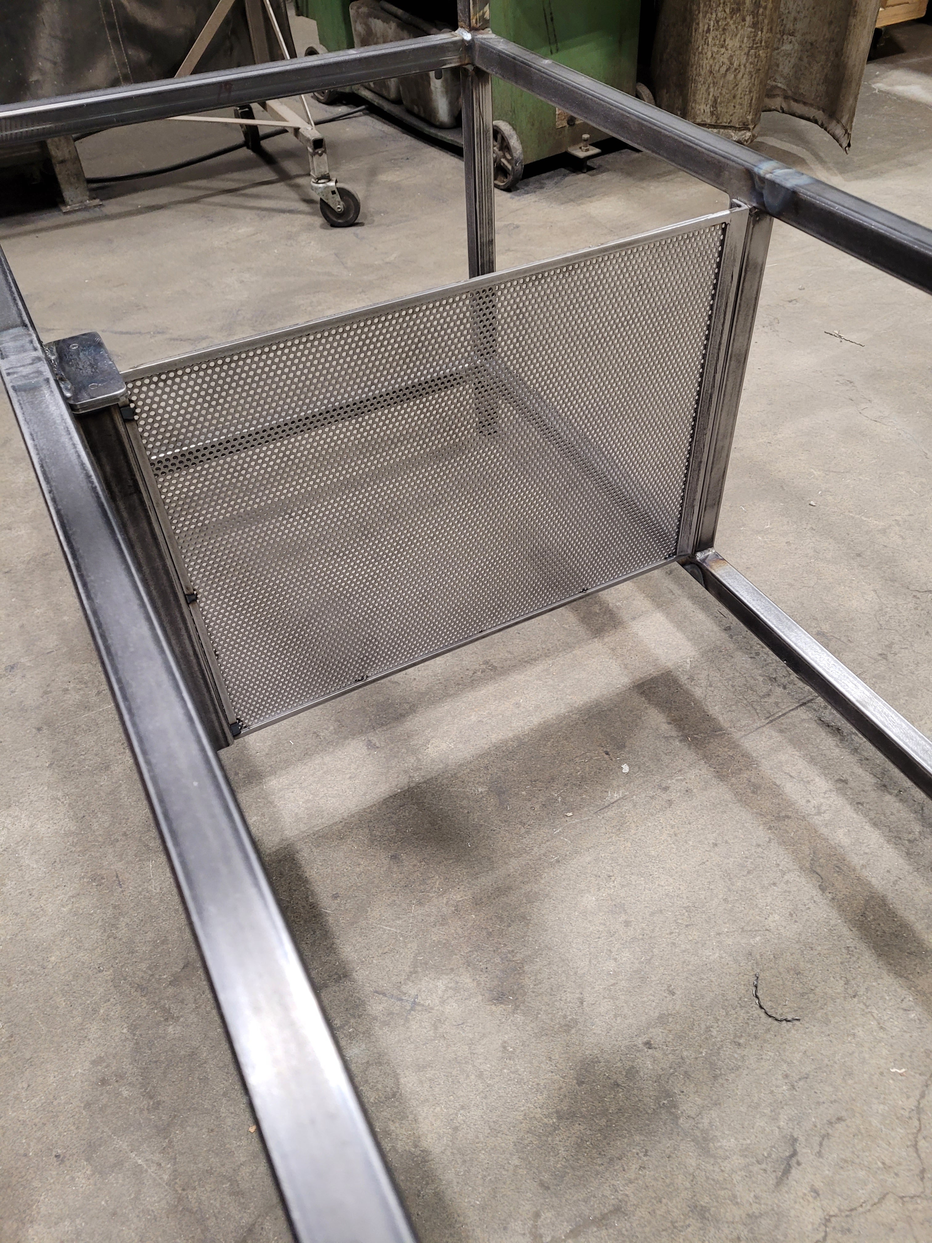 A stainless steel mesh shelf or divider mounted on a metal frame, photographed in an industrial workshop setting with a concrete floor.