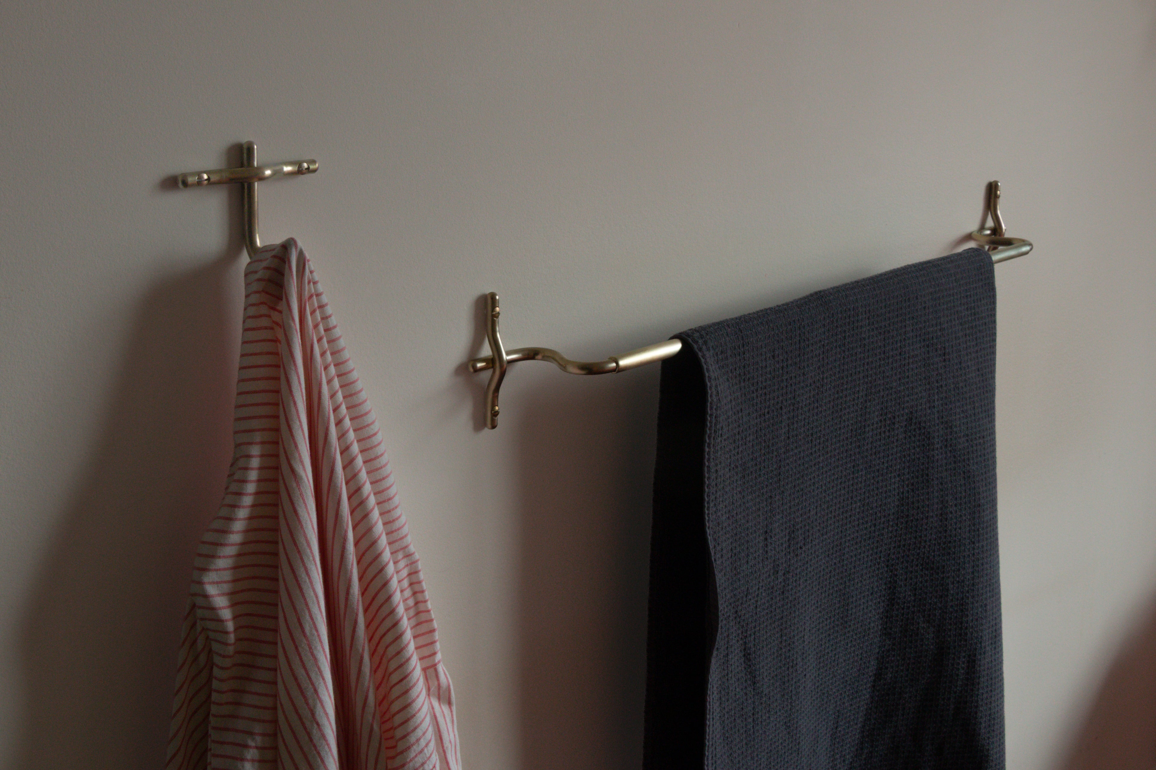 A brass wall-mounted towel bar holding a dark gray textured towel, with a striped pink and white towel hanging from a matching hook on an off-white wall.