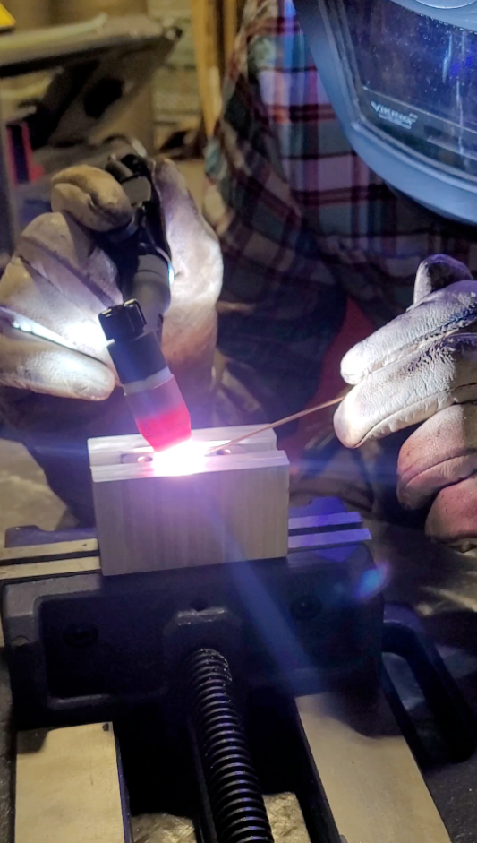 A close-up action shot of a person wearing protective welding gloves and a welding helmet, actively welding a small metal block held in a clamp. The bright welding arc illuminates the scene, set against a workshop background.